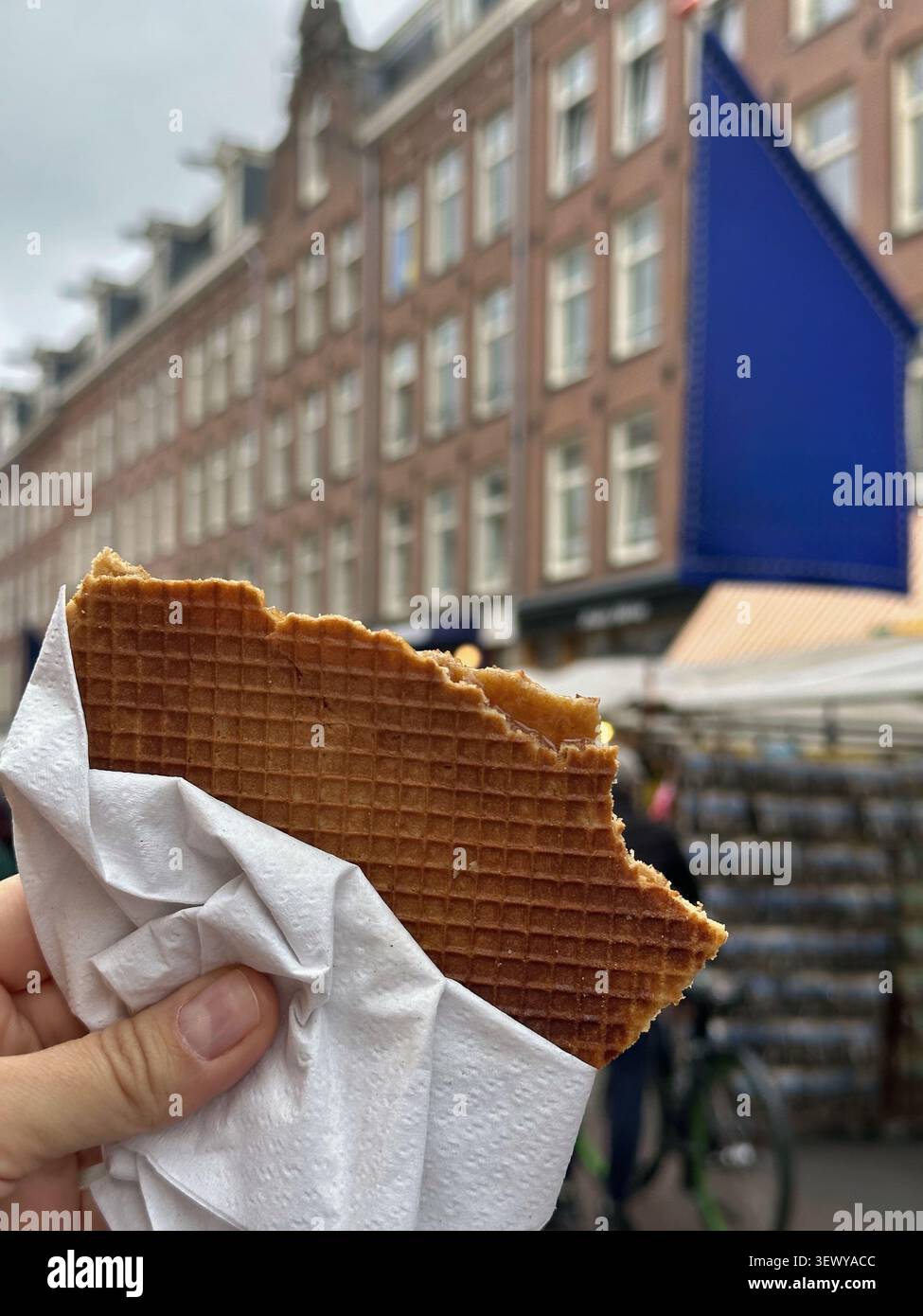 Traditionelle niederländische Stroopwafel in der Hand auf einem Straßenmarkt in amsterdam. Eine Hand hält eine köstliche gebissene niederländische Stroopwafel, die in eine weiße Napki gewickelt ist - Smartphone-aufgenommenes Stockfoto