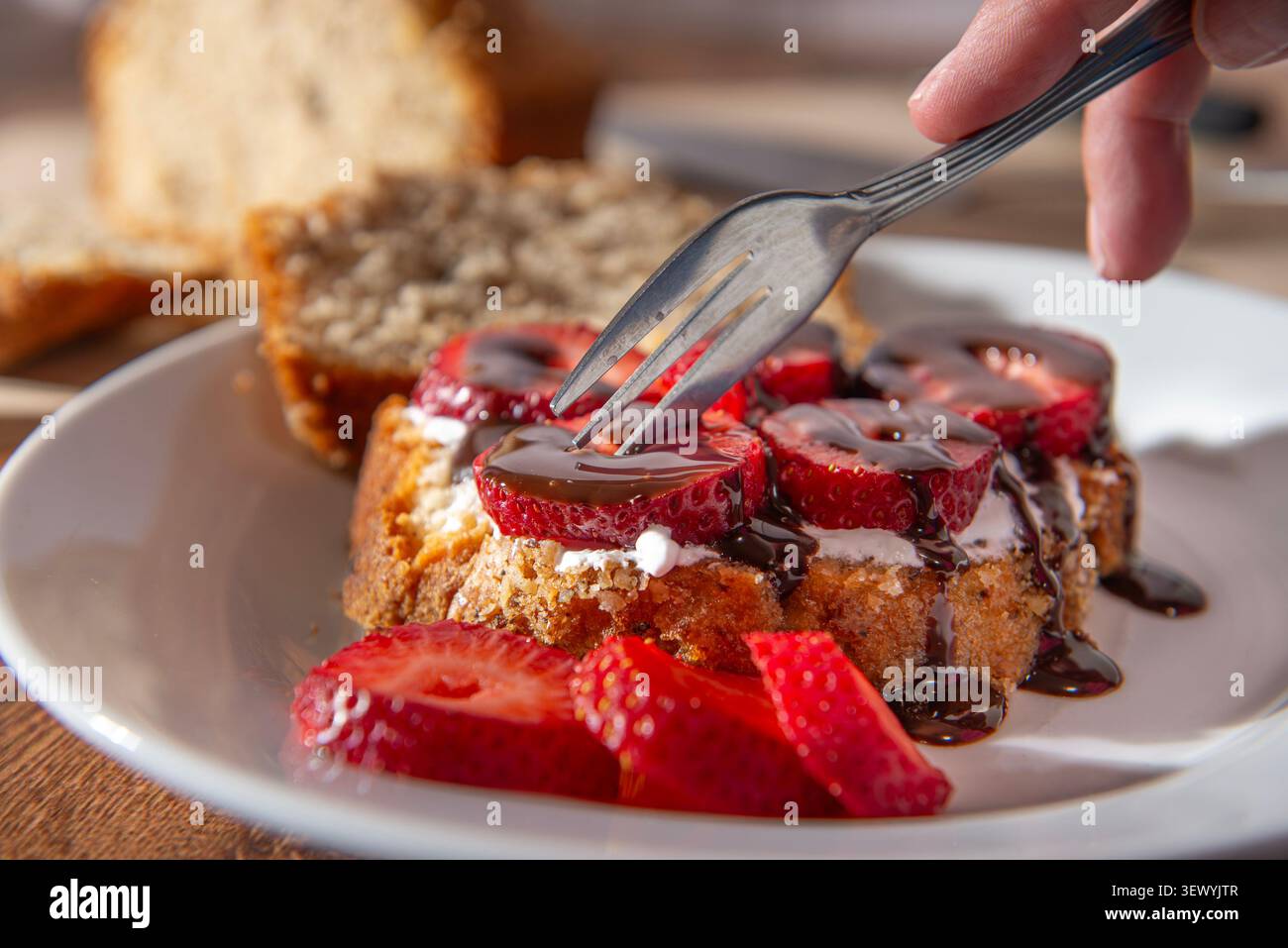 Nahaufnahme, Seitenaufnahme einer Frauenhand mit einer Gabel, die in ein Nusskuchen-Dessert mit Schlagsahne und Erdbeeren mit Schokoladenüberzug geschnitten wird. Stockfoto
