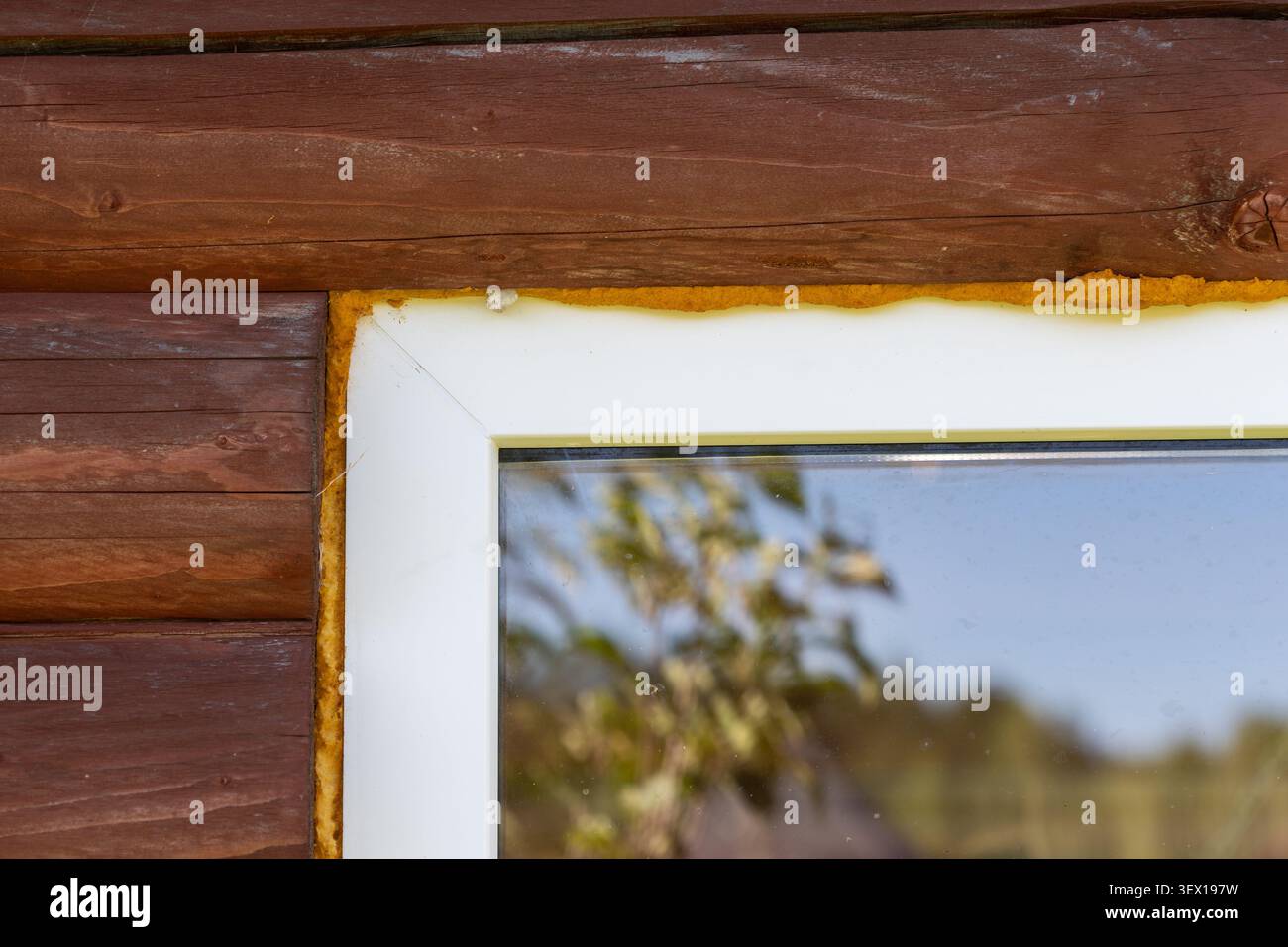 Gelber Polyurethanschaum versiegelt eine weiße Fensterrahmenecke an der Außenwand eines braunen Holzholzhauses mit Reflexion in Glas. Stockfoto