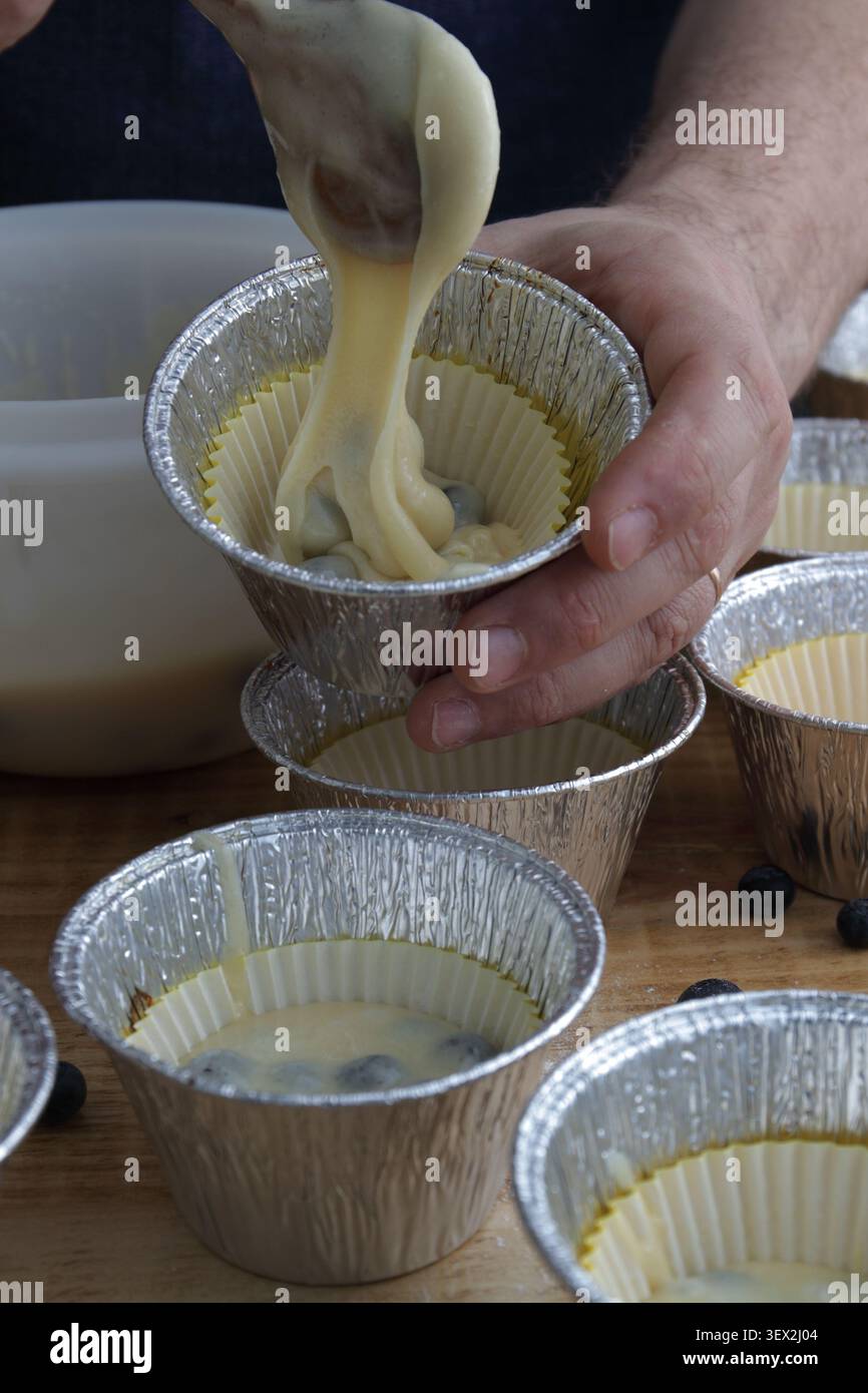 Hausgemachter Blaubeeren-Muffin-Teig, der in Einweg-Backformen in Papierbecher gegossen wird. Nahaufnahme des Backprozesses vor den Muffins Stockfoto