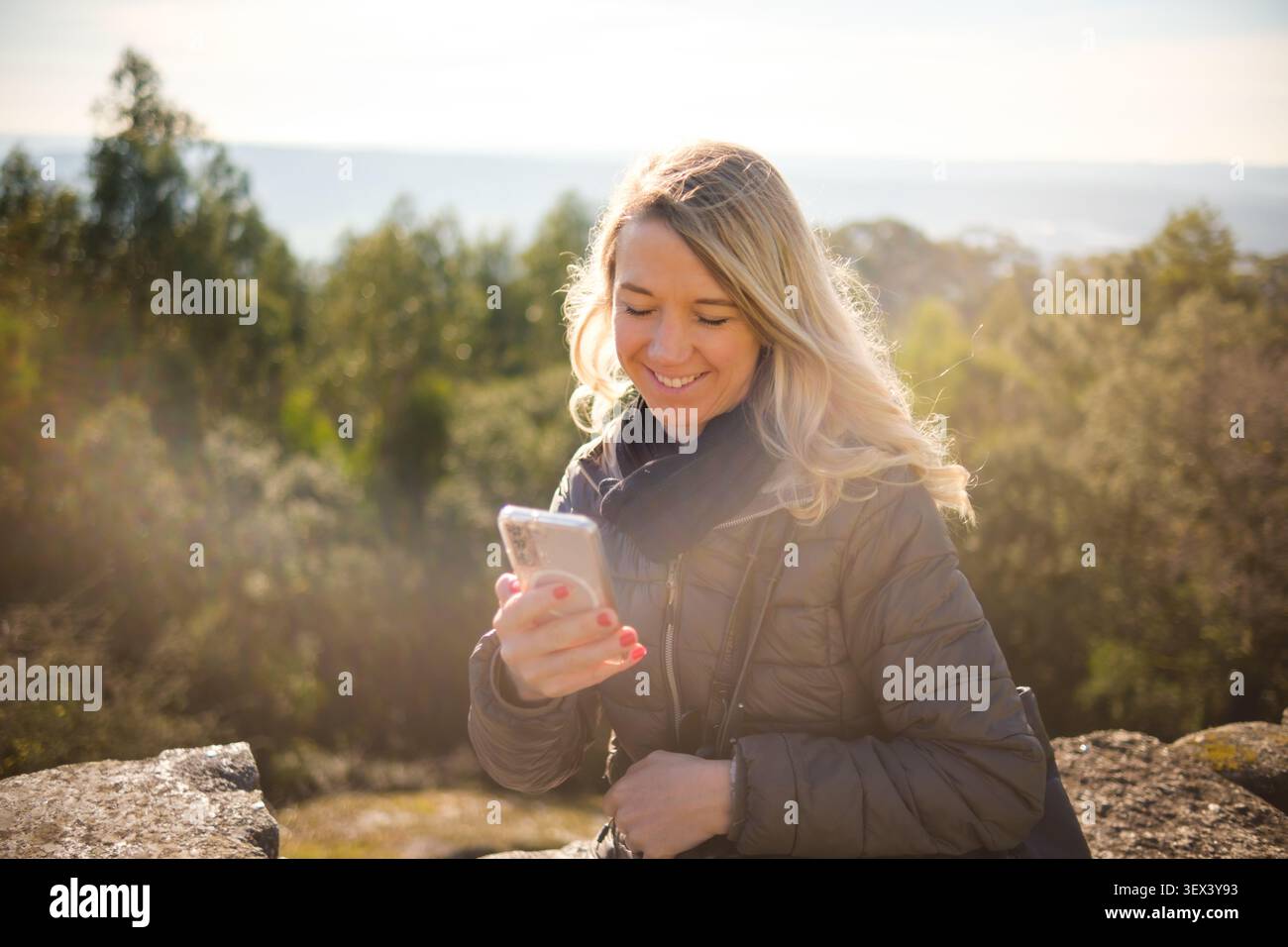 Blonde Frau genießt sonnigen Herbsttag, überprüft Handy in der Naturlandschaft Stockfoto