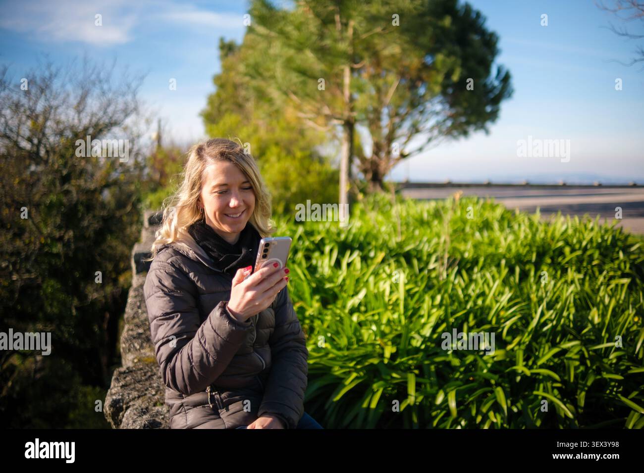 Frau lächelt und schreibt per Smartphone, während sie sich in einer wunderschönen Landschaft im Freien entspannt Stockfoto