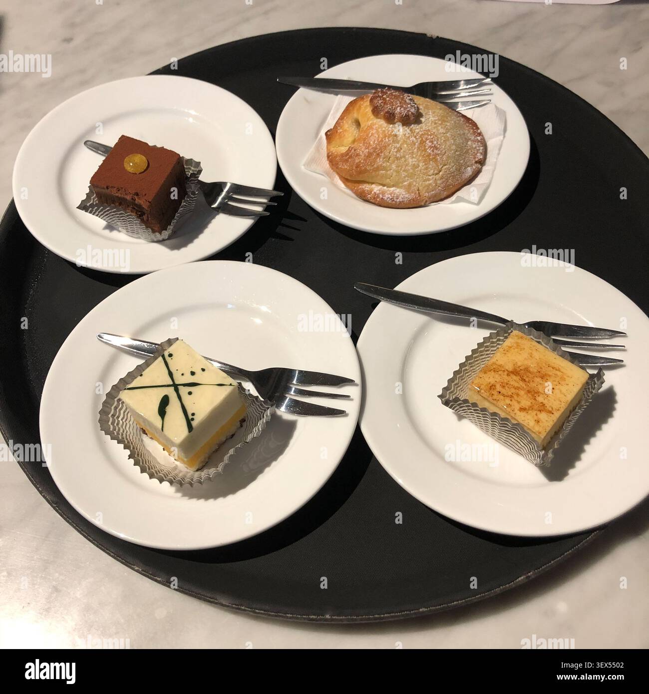 Ein Blick von oben auf vier verschiedene Miniatur-Desserts, die auf kleinen weißen Tellern auf einem runden schwarzen Tablett angeordnet sind und kulinarische Genüsse wecken. Stockfoto