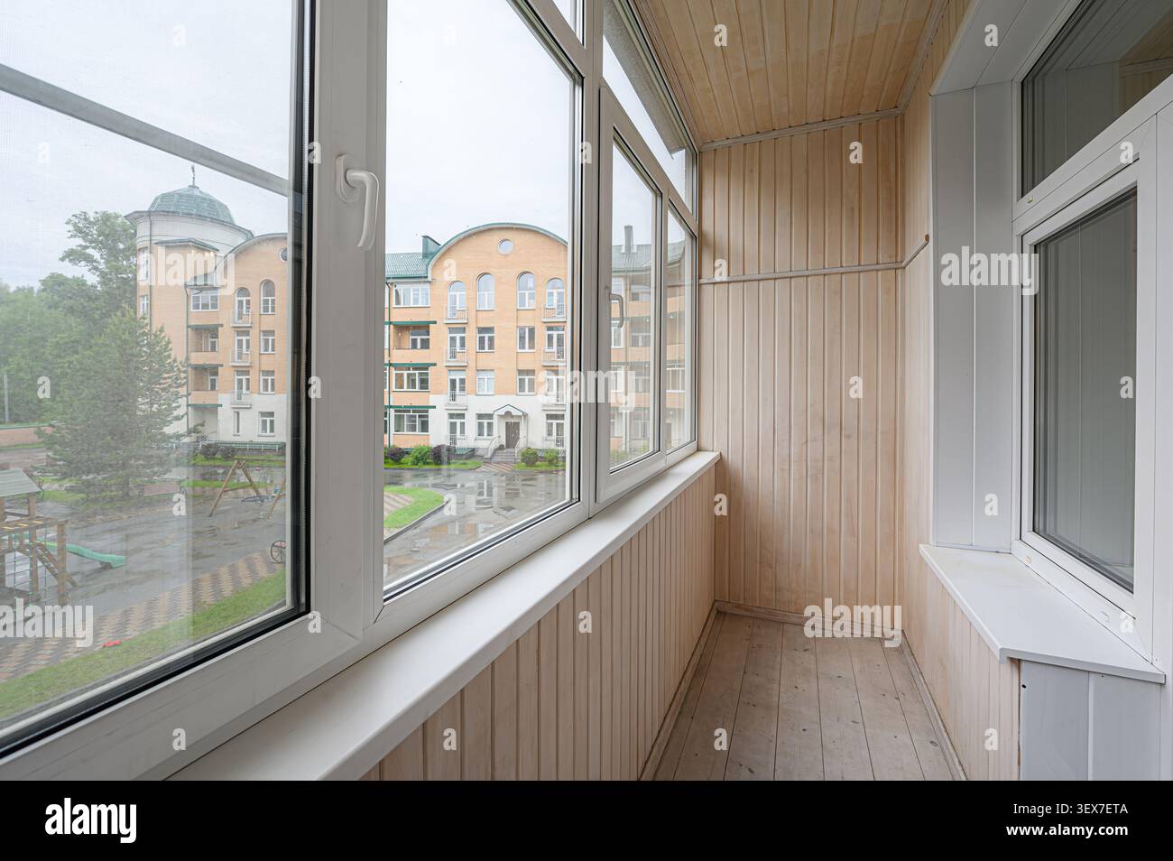 Heller Balkon mit heller Holztäfelung und großen Fenstern, die eine nasse Stadtlandschaft mit Gebäude und Spielplatz darstellen Stockfoto