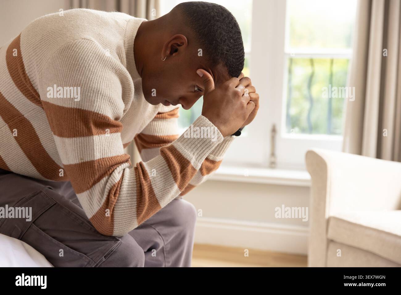 Erwachsener Mann, der auf dem Bett im Schlafzimmer sitzt, sich nach vorne lehnt, einen gestreiften Pullover und eine Uhr trägt, Kopierraum Stockfoto