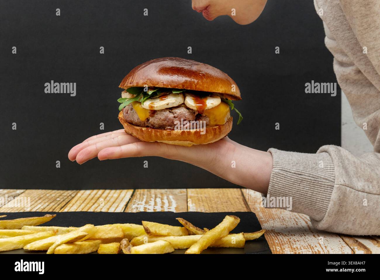 Person mit Gourmet Burger mit geschmolzenem Käse, frischem Arugula und gegrilltem Halloumi auf einem Brioche-Brötchen, serviert mit rustikalen Pommes frites Stockfoto