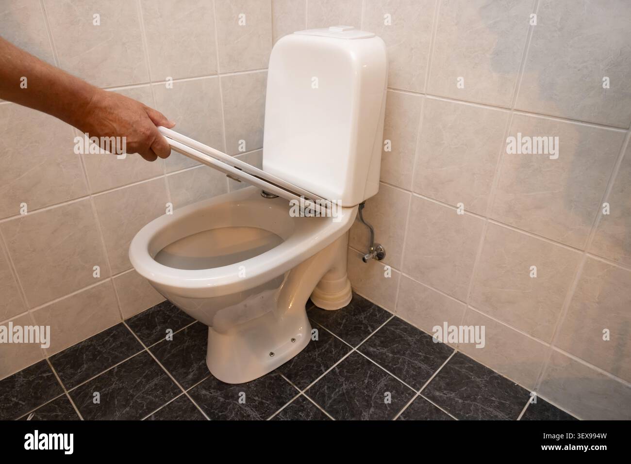 Reinigt die Toilettenschüssel mit einer Bürste von Hand. Weiße Porzellanbefestigung an Fliesenwänden. Dunkle Bodenfliesen stehen im Kontrast zu hellen Wänden. Der Benutzer konzentriert sich auf Sanitat Stockfoto