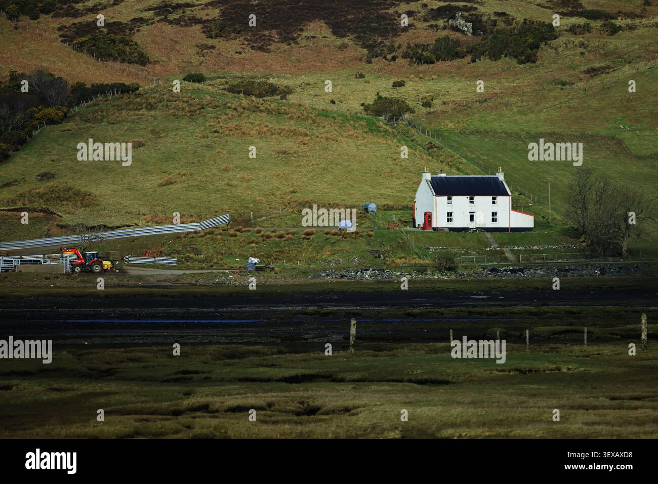 Roadtrip-Landschaft auf der Isle of Skye in den schottischen Highlands, Schottland, zeigt ein weißes Cottage neben einer dunklen Straße mit grünen Hügeln und Gebäuden am Straßenrand. Stockfoto