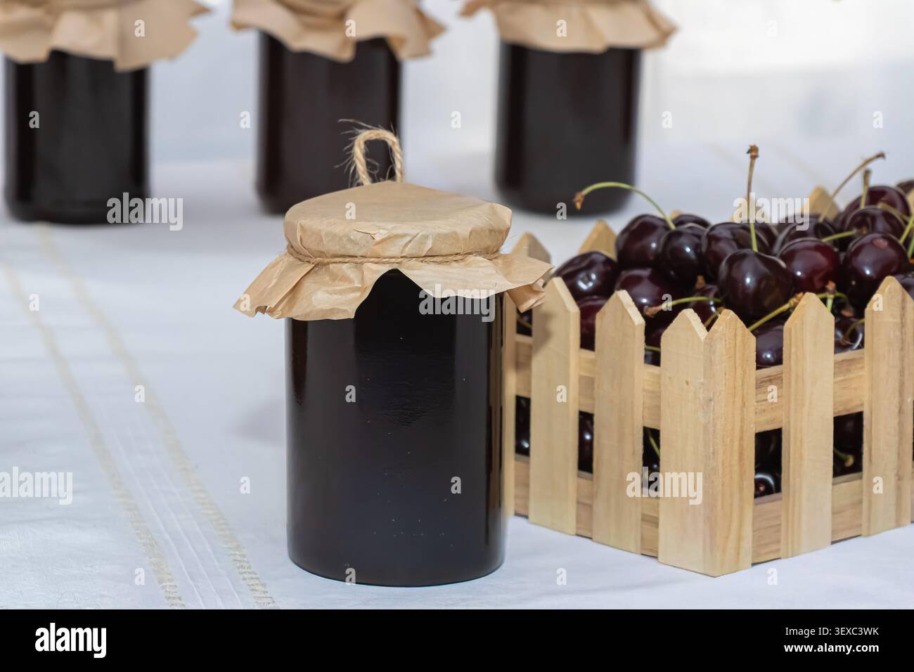 Dunkle hausgemachte Kirschmarmelade in einem Glasgefäß mit Kraftpapierüberzug neben Reifen Kirschen in einer rustikalen Holzkiste, fotografiert als sauberes Stillleben Stockfoto