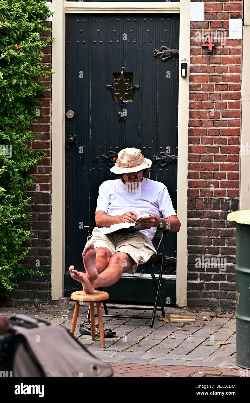 Barfußer Mann liest ein Buch vor einem traditionellen niederländischen Haus, während in der Nähe ein lokales Straßenevent stattfindet Stockfoto