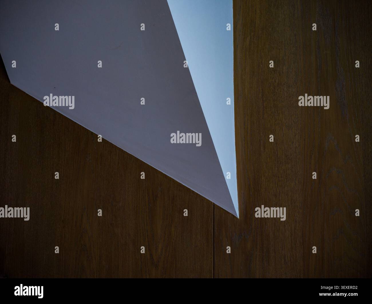 Abstraktes architektonisches Detail mit einem scharfen geometrischen weißen Dreieckselement, das in eine warme braune holzgetäfelte Wand geschnitten ist. Stockfoto