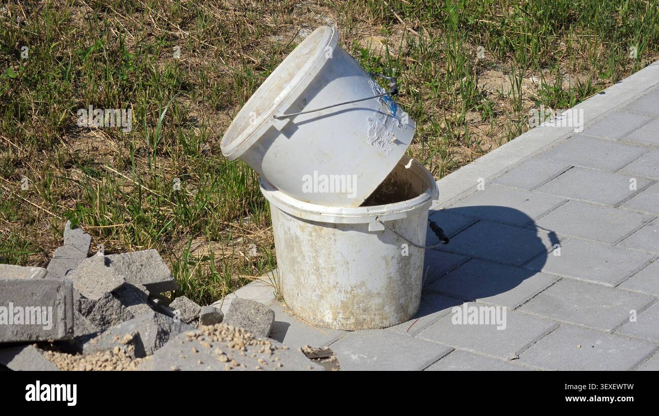 Ein Paar abgenutzte weiße Plastikeimer, die mit getrockneten Mörtelrückständen verschmiert sind, stehen ineinander verschachtelt auf einem grauen, verzahnten Steinplatten-Fußweg Stockfoto