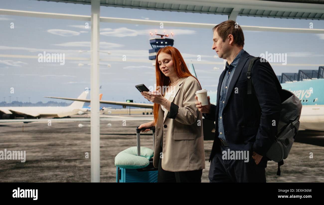 Geschäftsleute laufen im Flughafenterminal, besprechen arbeitsbezogene Aufgaben, verwenden Mobiltelefon. Digitale Nomaden, die eine Kaffeetasse halten, unterhalten sich am Flughafen über ein Smartphone Stockfoto