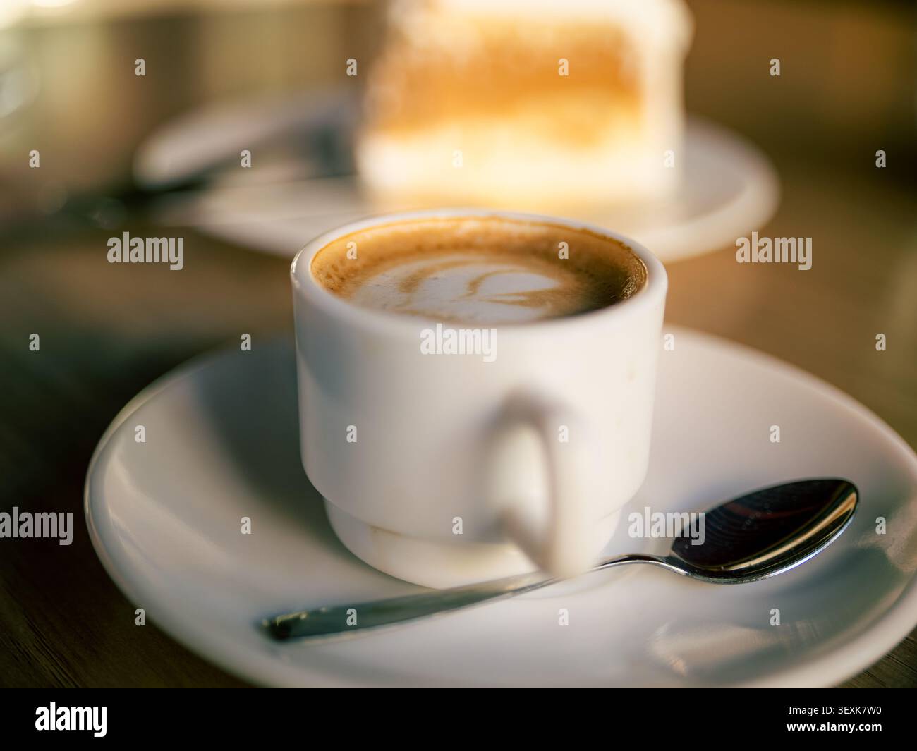 Die weiße Keramikschale hält Kaffee mit Latte Art. Silberlöffel liegt auf weißer Untertasse. Unscharfe Kuchenscheibe wird im Hintergrund mit weichem Fokus angezeigt. Warmes Licht Stockfoto