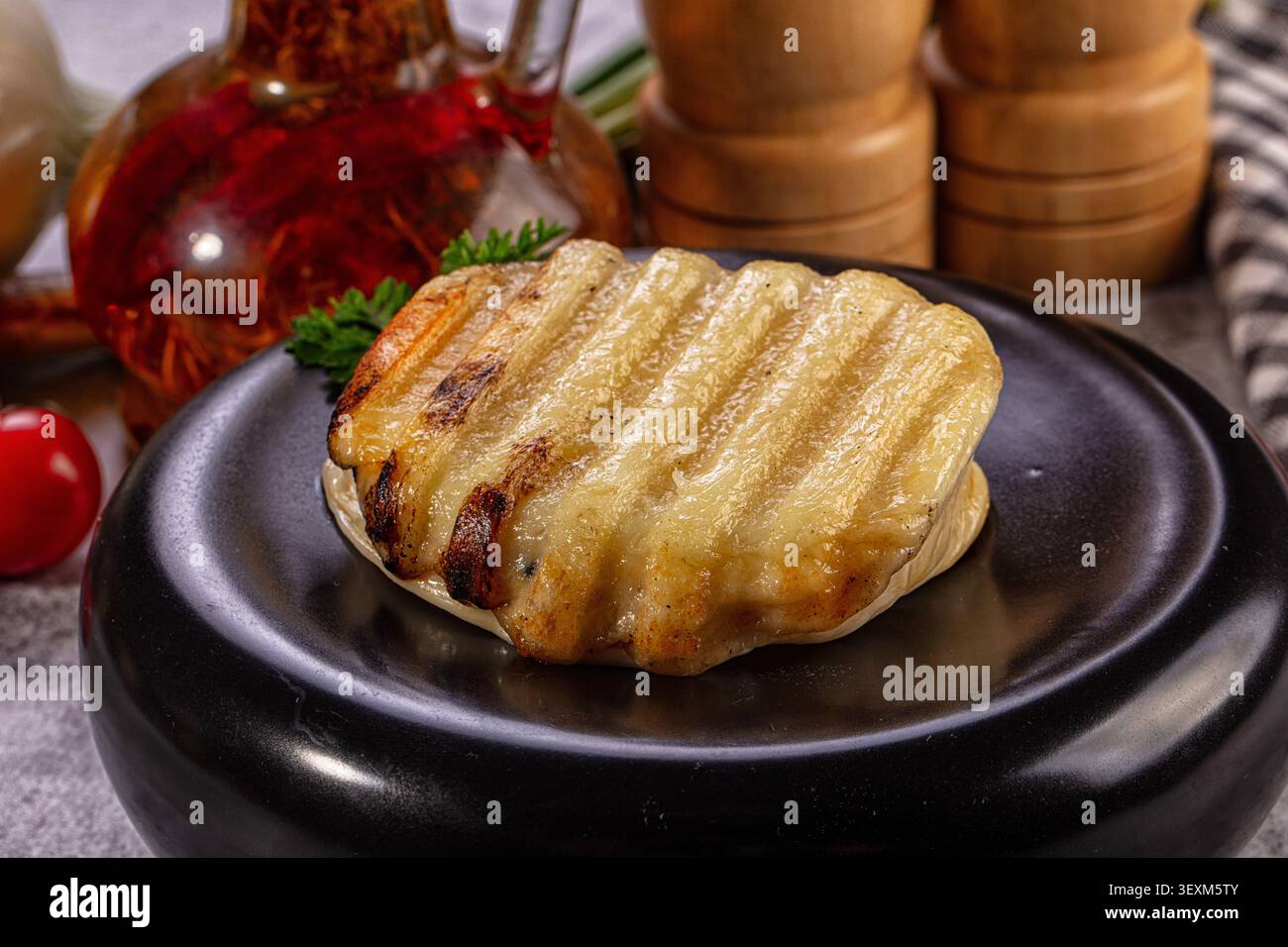 Goldbraune gegrillte halloumi-Käsescheibe mit Grillmarkierungen auf schwarzem Teller, garniert mit Petersilie, roter Ölflasche und hölzernen Salzmühlen im Hintergrund Stockfoto