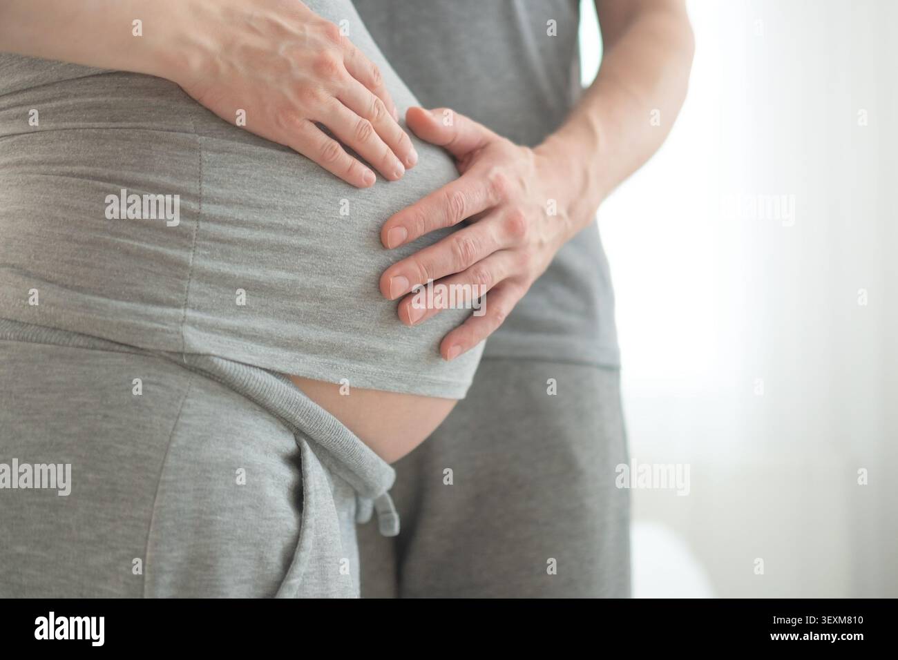 Nahaufnahme einer kräftigen stützenden Hand mit festem Griff, die den schwangeren Bauch in pflegender Schutzumarmung umhüllt. Die erwartete Mutter und der Mann teilen sich einen zärtlichen Moment in einem minimalen Studio mit sauberem weißem Hintergrund. Stockfoto