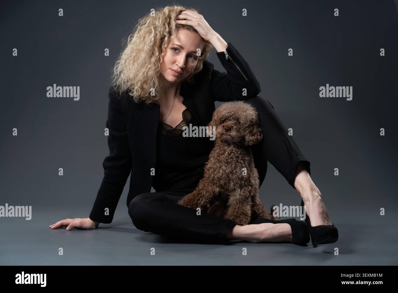 Eine junge Frau sitzt auf dem Boden eines dunklen Ateliers mit einem braunen Spielzeugpudel, der sich in ihre Seite lehnt. Ihre entspannte Haltung sorgt für einen stilvollen und modernen Look Stockfoto