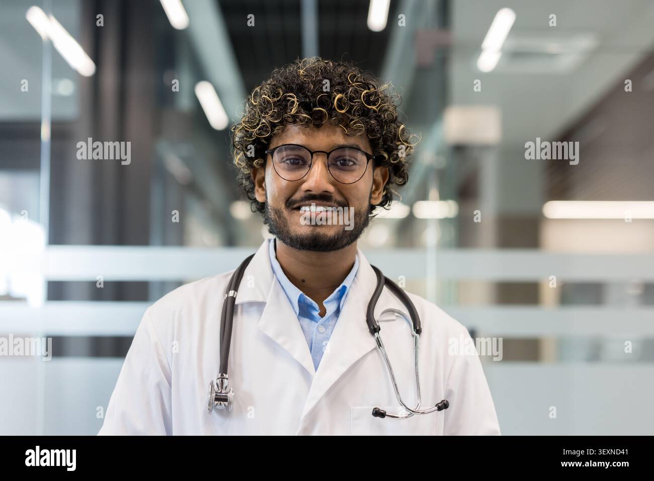 Junger indischer Arzt mit lockigen Haaren und Brille. Mit weißem Labormantel und Stethoskop. Selbstbewusst lächelnd im Krankenhaus oder in der Klinik. Vertretung von professionellen Gesundheitsdiensten und medizinischen Dienstleistungen Stockfoto