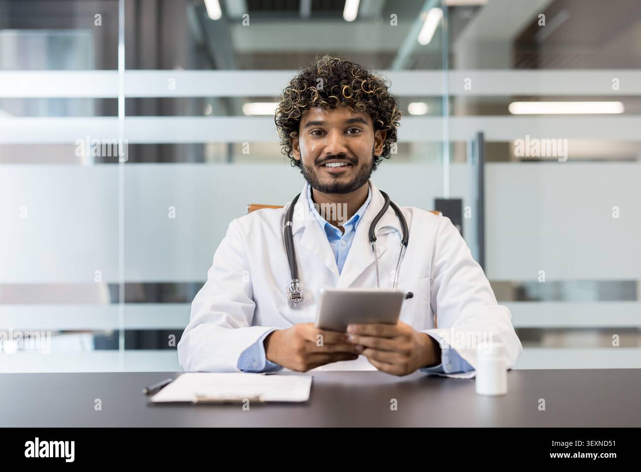 Junger männlicher Arzt mit lockigen Haaren und einem Stethoskop, der einen weißen Labormantel trägt, an einem Schreibtisch sitzt und ein digitales Tablet hält, das moderne Kommunikation und Technologie im Gesundheitswesen repräsentiert Stockfoto