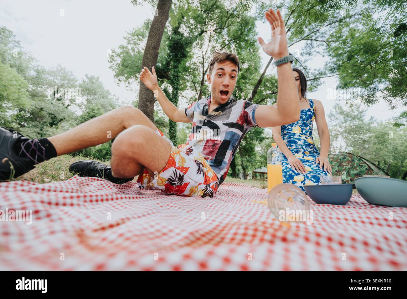 Überraschter junger Mann, der auf eine Picknickdecke fällt, während ein Paar im Sommer ein Picknick im Park genießt Stockfoto