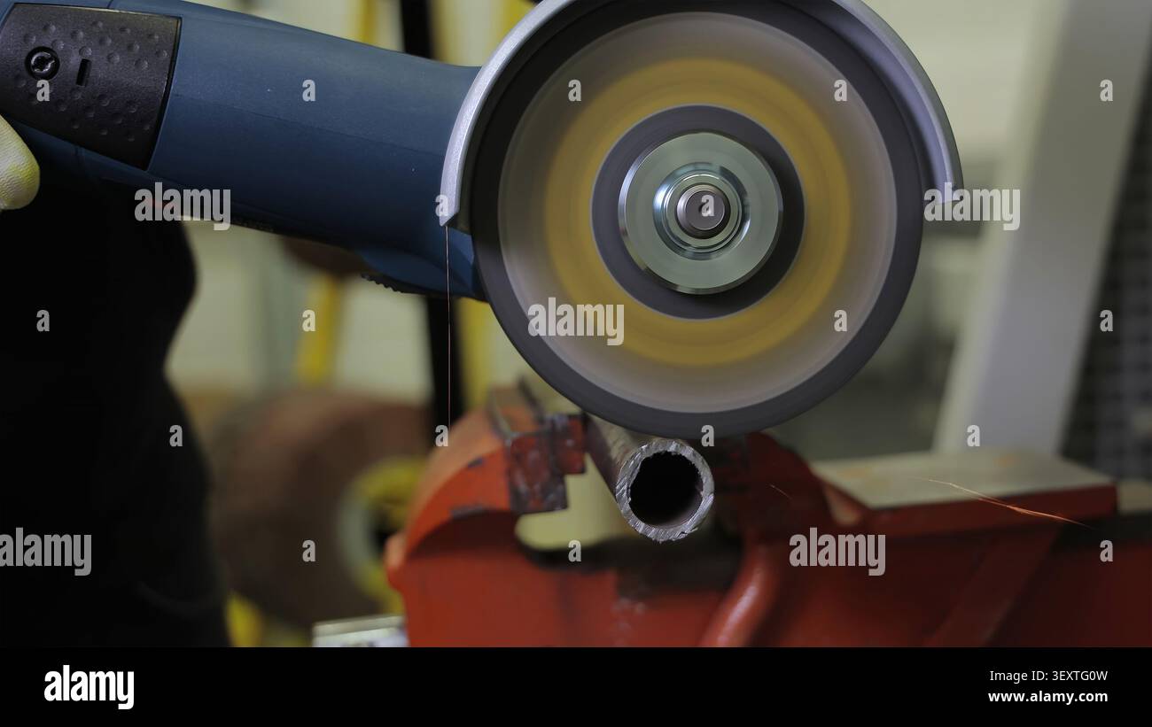Winkelschleifer, der Metallrohre in der Werkstatt schneidet. Der Winkelschleifer mit einer drehenden Schleifscheibe schneidet ein Metallrohr, das in einem roten Schraubstock gehalten wird, und erzeugt so Stockfoto