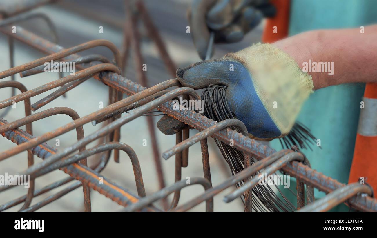 Arbeiter Mit Handschuhen Binden Rebar Mit Stahldraht. Bauarbeiter mit Handschuhen binden rostigen Bewehrungsstahl mit Stahldraht an einem Verstärkungskäfig mit Zangen und Stockfoto