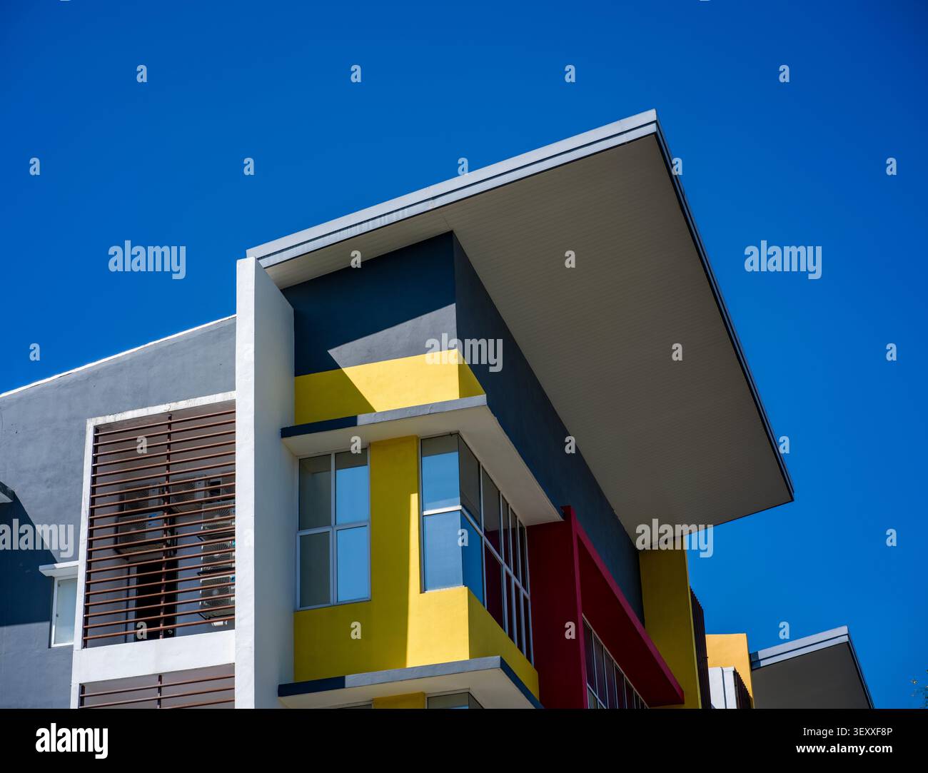 Flache Aufnahme einer modernen Gebäudefassade mit scharfen geometrischen Linien, hellen gelben und roten Akzenten und einem überhängenden Dach unter einem leuchtenden blau Stockfoto