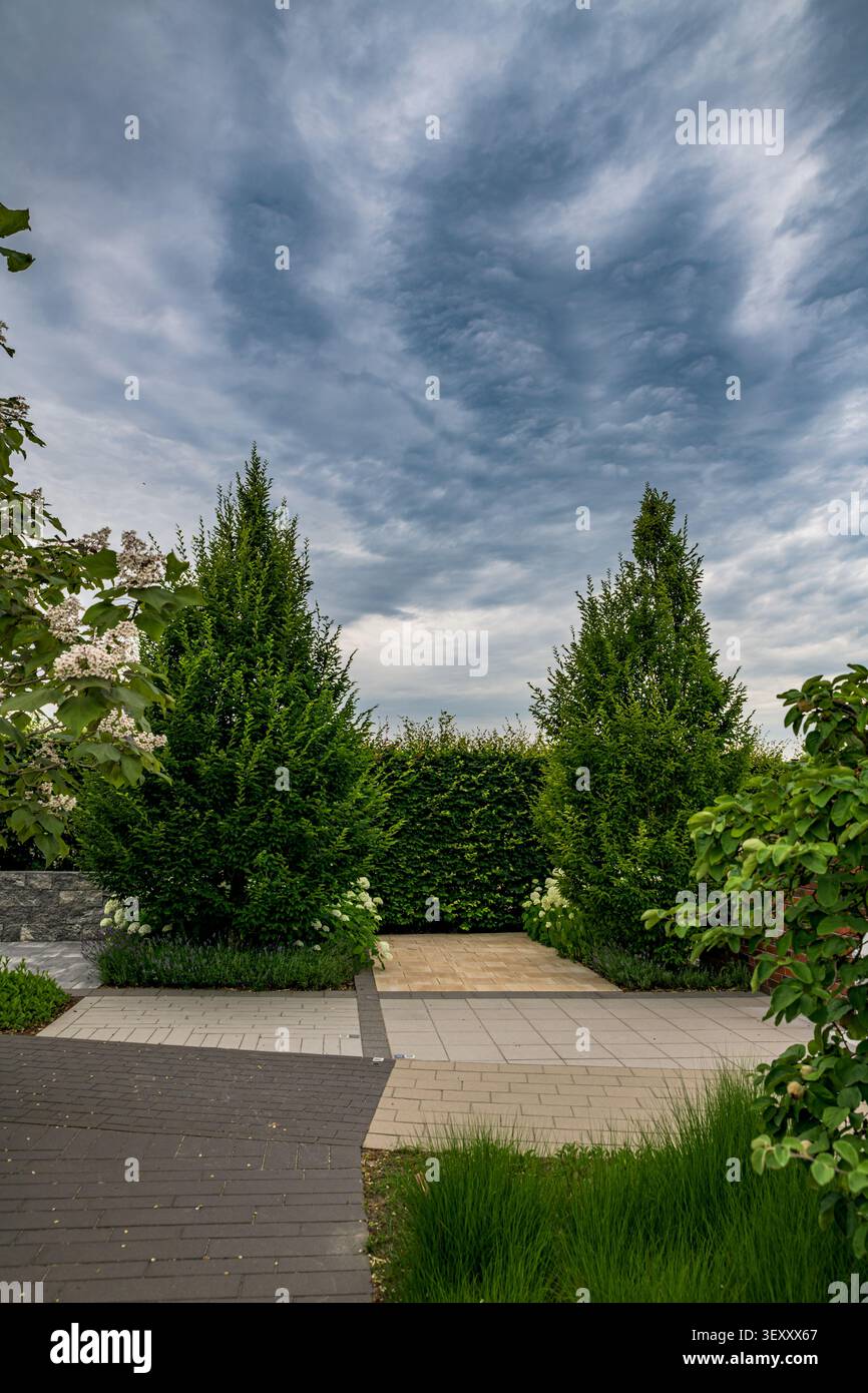 Symmetrisch gestaltete Gartenzone mit zwei pyramidal geformten Hornbeum Bäumen und stein gepflasterten Wegen unter einem dramatischen bewölkten Himmel Stockfoto