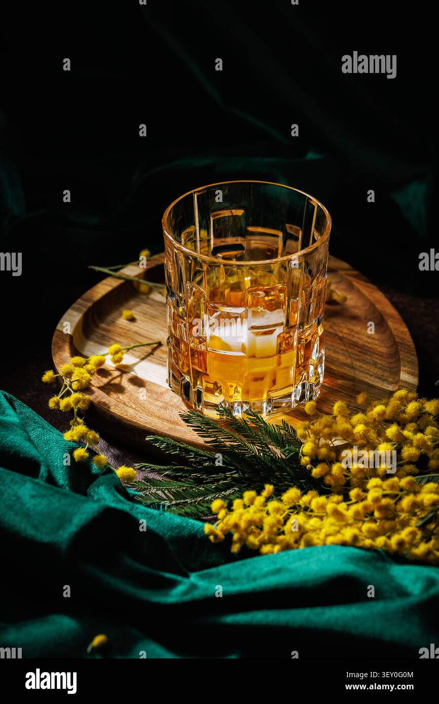 Kristall-Whiskeyglas mit Eis auf Holztablett mit Mimosenblüten für ein Frühlingsgetränk Stockfoto