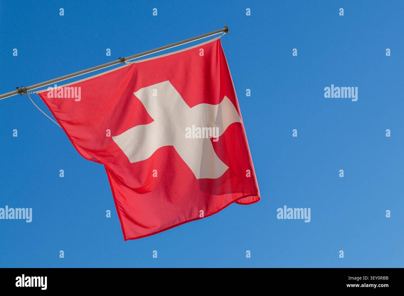 Niedrigwinkelaufnahme der offiziellen Schweizer Nationalflagge mit dem weißen Kreuz auf rotem Grund, die im Wind weht. Minimalistische Komposition unter einem blauen Himmel Stockfoto