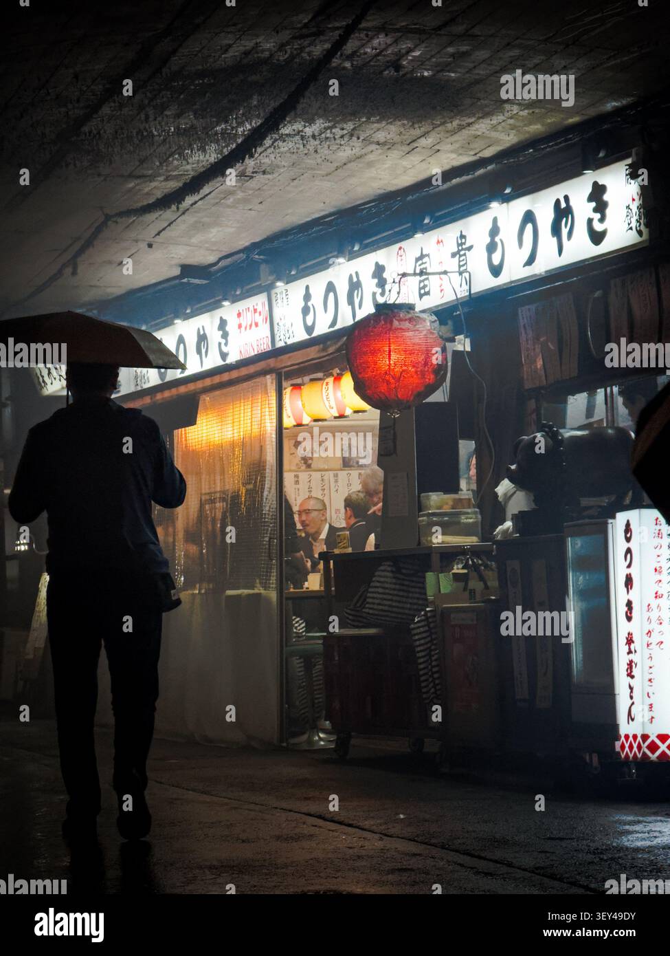 Stimmungsvoller Nachtblick auf ein traditionelles Izakaya Restaurant in Tokio, Japan. Silhouette einer Person, die in einer regnerischen Nacht mit einem Regenschirm läuft. - Smartphone-aufgenommenes Stockfoto