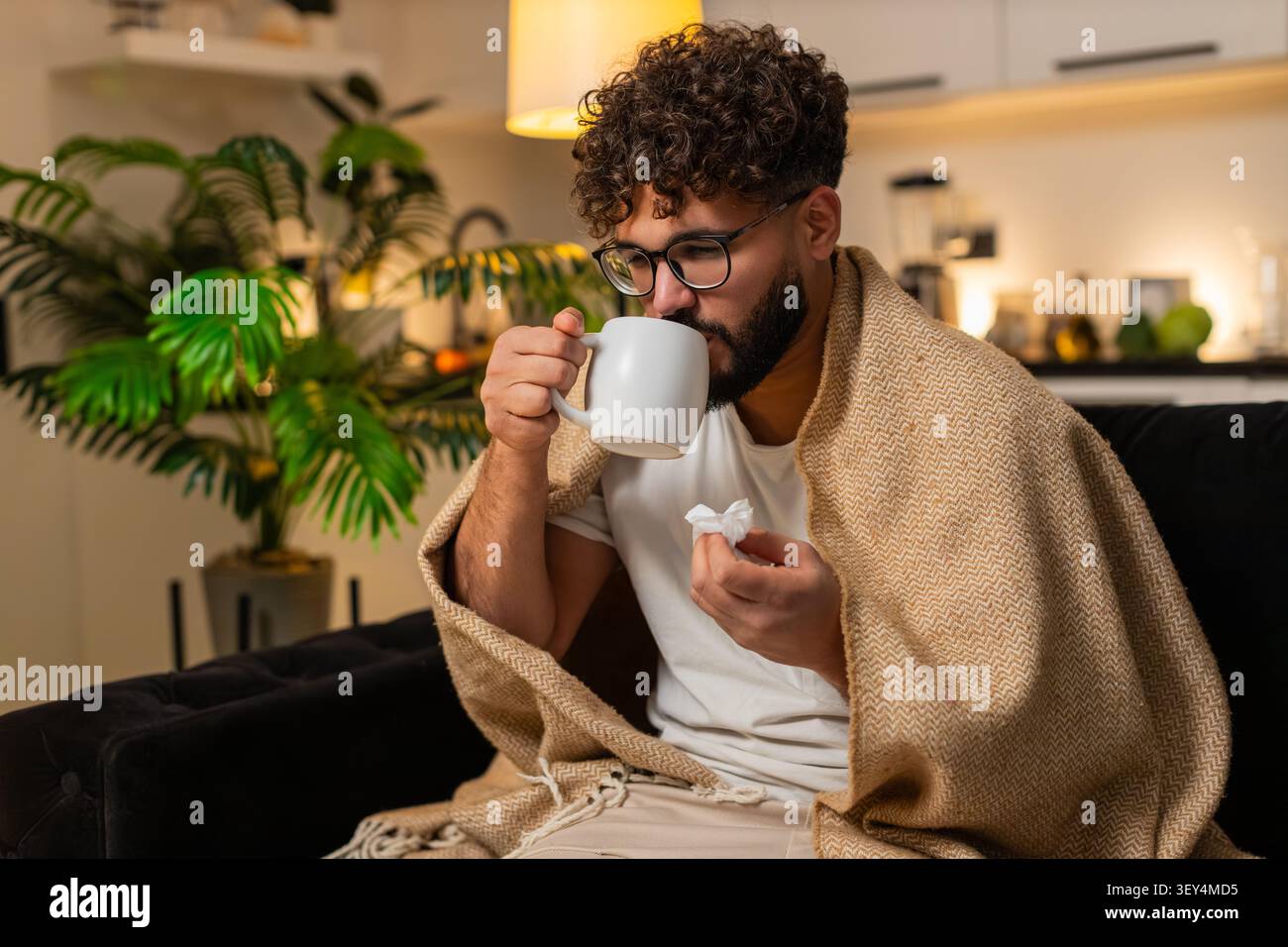 Arabian Mann zu Hause fühlt Halsschmerzen hält Tasse heißen Tee mit Medizin nimmt langsamen Schluck verwendet Gewebe. Ägyptischer Kerl auf Sofa durchsucht Arzt auf Smartphon Stockfoto