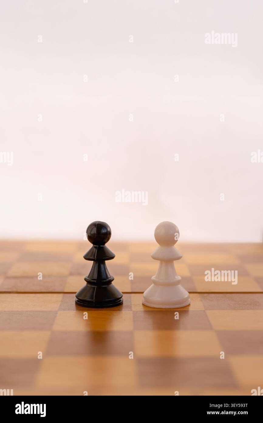 Eine vertikale Nahaufnahme eines einzelnen schwarzen Bauern und eines weißen Bauern, der von Angesicht zu Angesicht auf einem hölzernen Schachbrett steht. Der Hintergrund ist sauber, hell, Stockfoto
