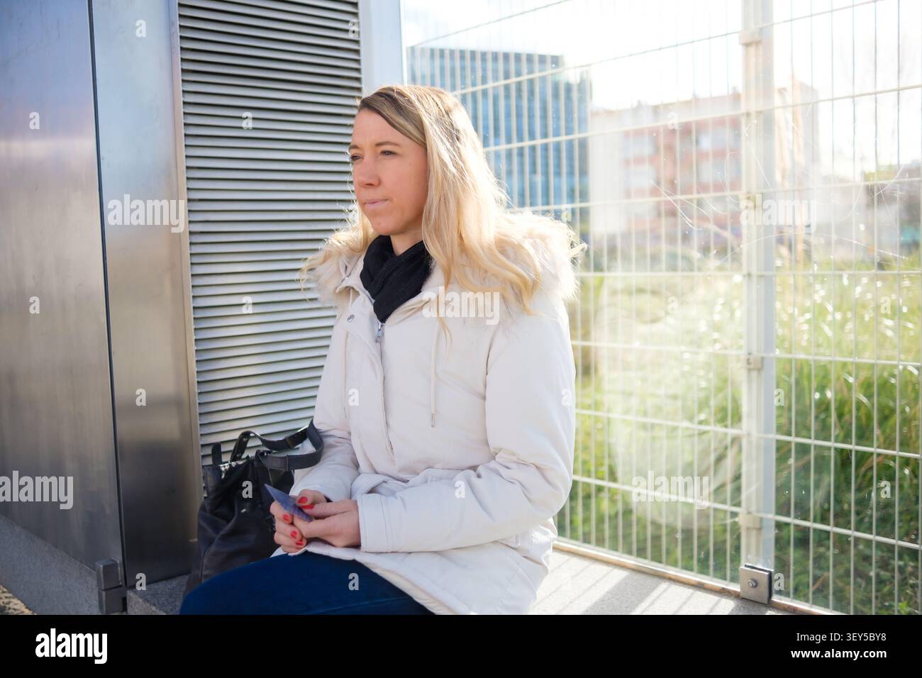 Blonde Frau, die in einer Winterjacke sitzt, eine Karte hält und in einer städtischen Umgebung nachdenkt Stockfoto
