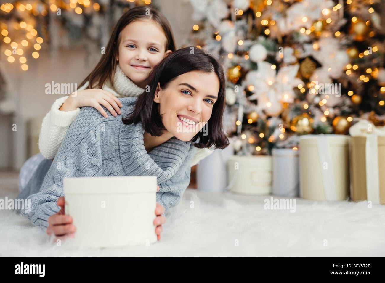Brünette lächelnde weibliche Erwachsene und ihr schönes entzückendes kleines Kind in weißem Pullover im Wohnzimmer, Geschenk halten, Vater und Ehemann mit bevorstehendem Neujahr oder Weihnachten gratulieren Stockfoto