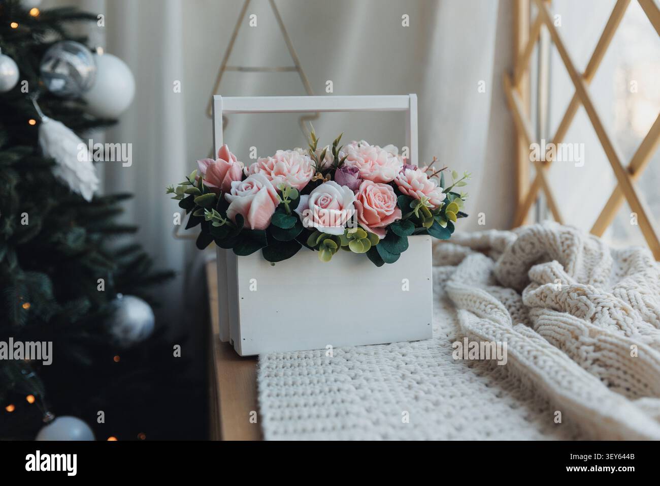 Weiche rosa Rosen ruhen in einer weißen Holzkiste. Kuschelige Strickdecken neben der festlichen Szene. Der Weihnachtsbaum leuchtet sanft mit silbernen Ornamenten. Wa Stockfoto