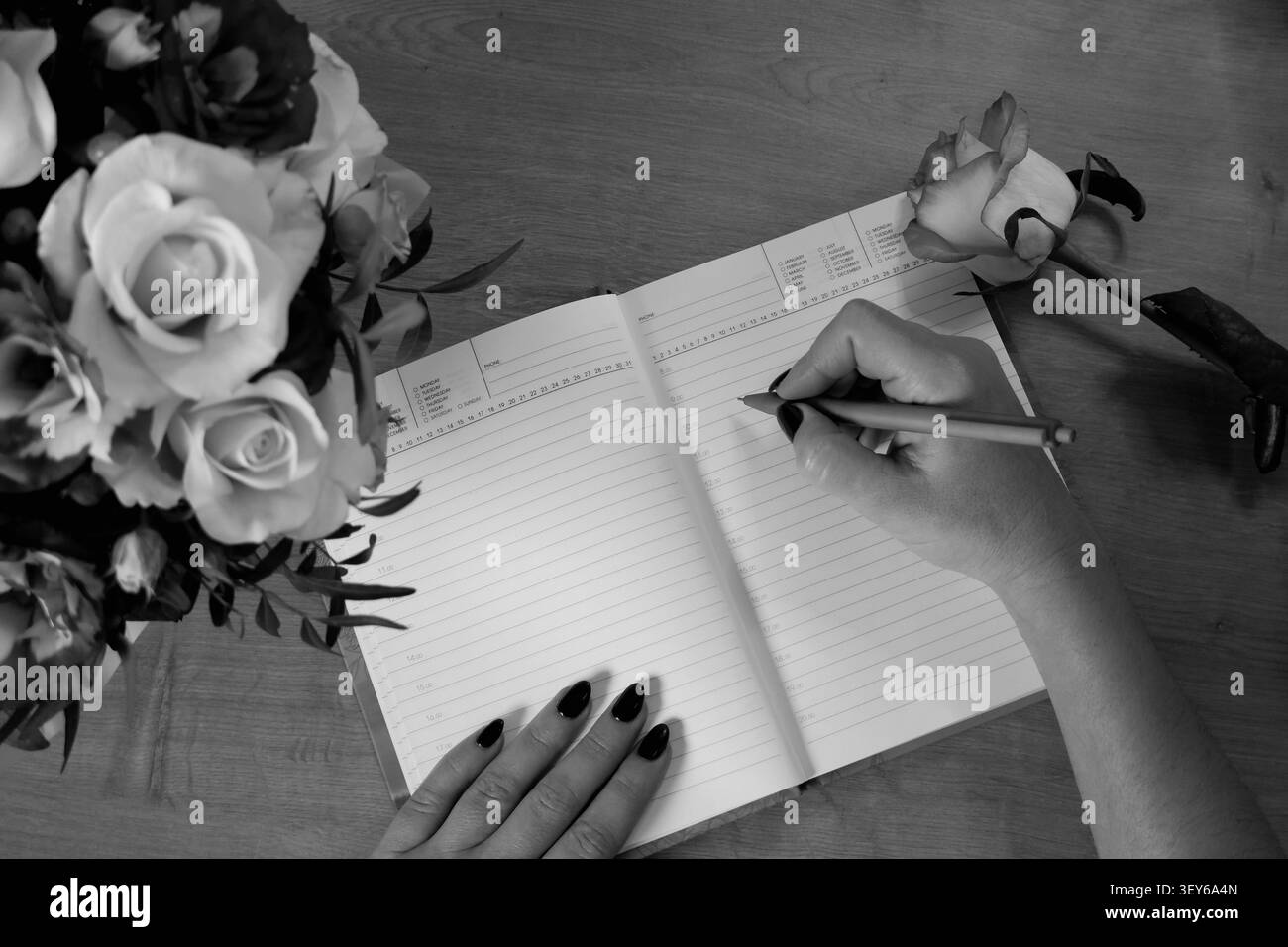 Weibliche Hände schreiben in offenem Notizbuch und Rosenstrauß auf alten Holztisch. Mit Stift planend, macht die Frau auf dem Tisch Einzug in ihr Tagebuch. Stockfoto
