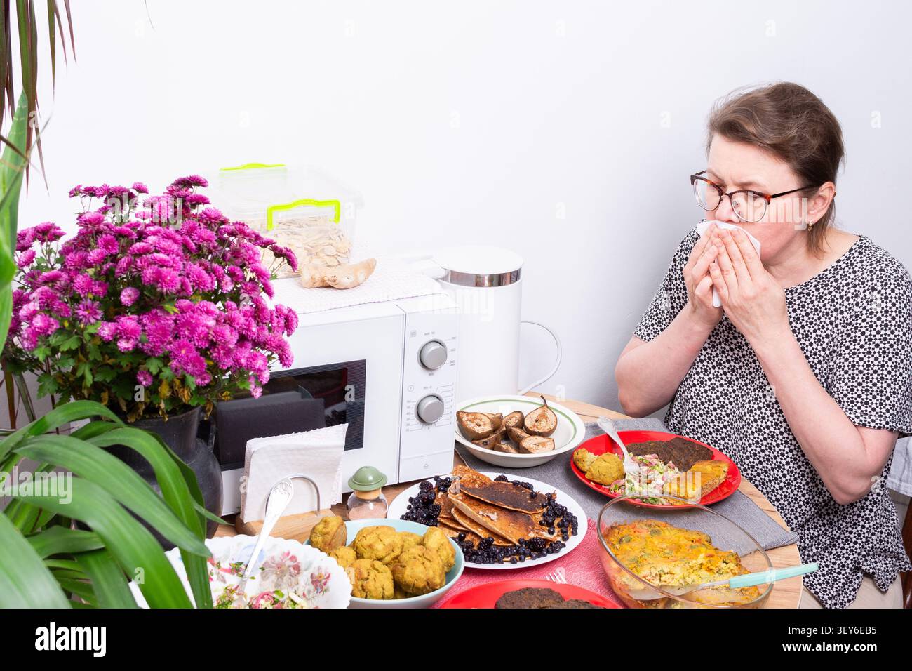 Eine Person sitzt an einem Küchentisch und isst. Um sie herum sind Teller mit verschiedenen hausgemachten Gerichten angeordnet. Blumen verleihen der Tischdekoration Farbe. Eine Mikrowelle steht in der Nähe Stockfoto