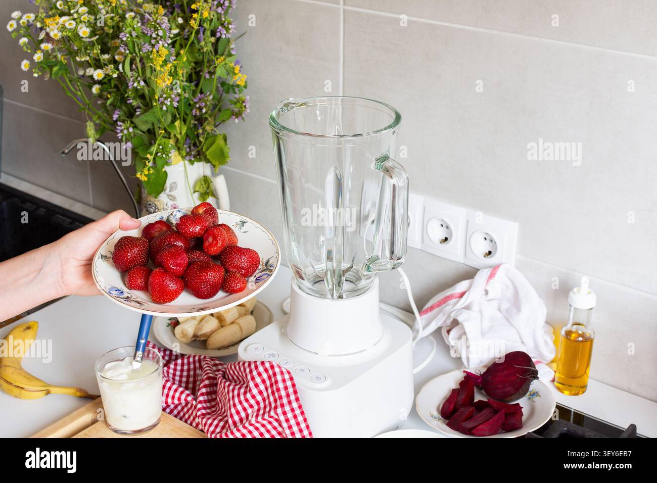 Eine Person hält einen Teller mit frischen Erdbeeren, während sie in einer Küche dabei ist, Eis zu machen. Zutaten wie Bananen und Rüben liegen ebenfalls auf der Arbeitsplatte zusammen mit einem Mixer. Stockfoto