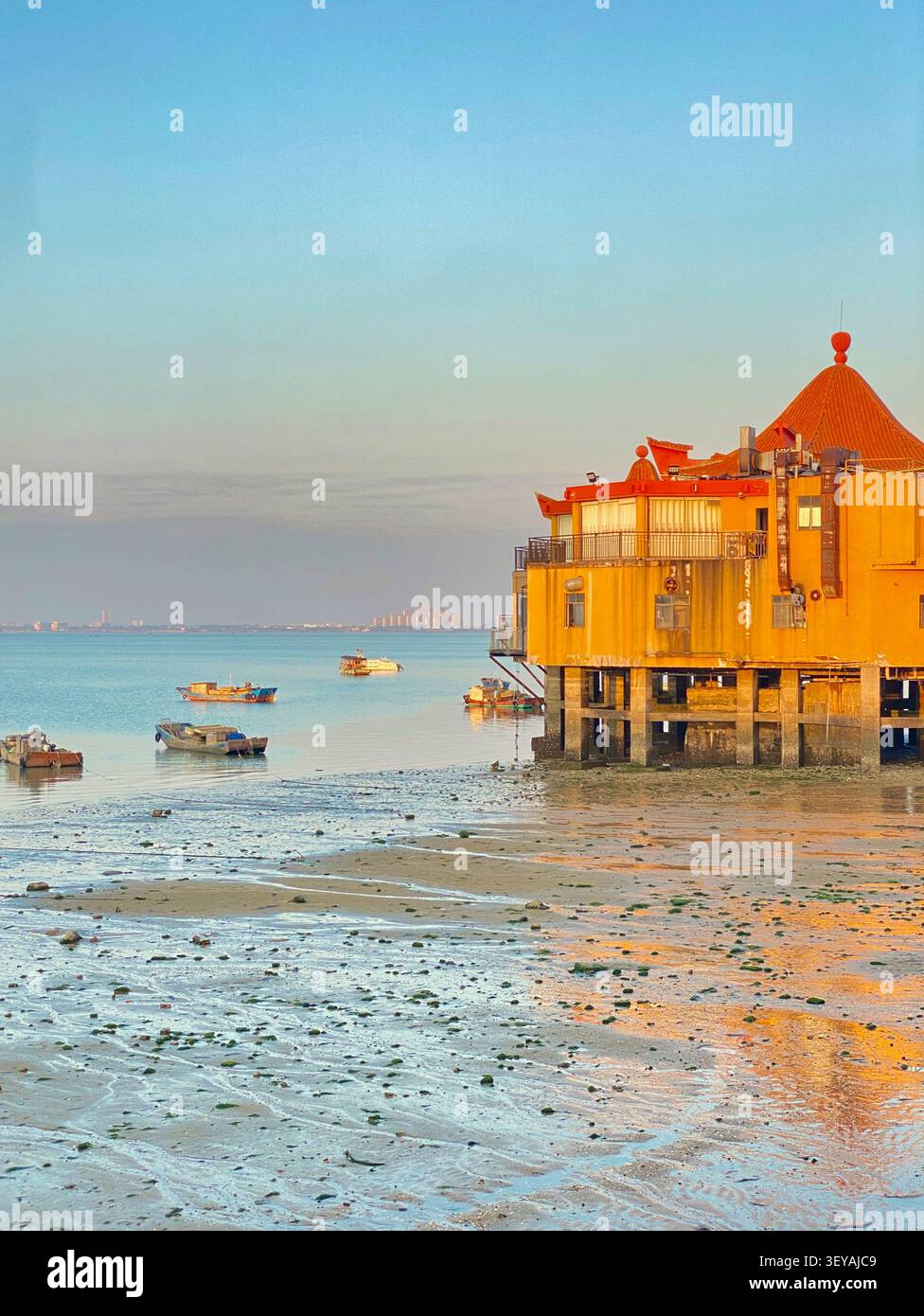 Gelbes chinesisches Stelzengebäude mit kunstvoll verziertem Pagodendach, das sich bei Sonnenuntergang in nassem Gezeitensand spiegelt, Fischerboote bei Ebbe, Küste Guangxi, Südchina - Smartphone-aufgenommenes Stockfoto