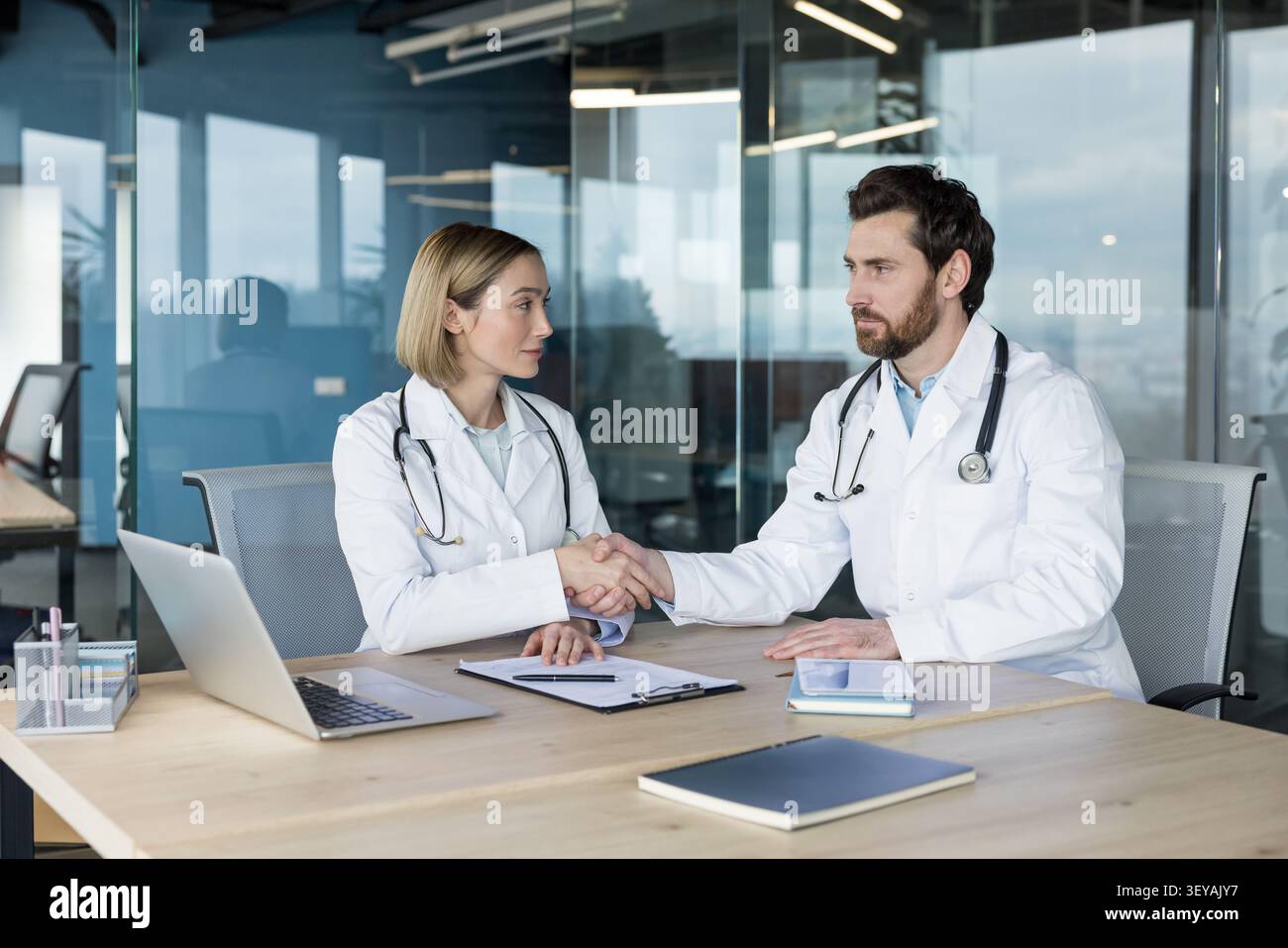 Zwei Ärzte. Ein Mann und eine Frau tragen Labormäntel und Stethoskope. Hände über einen Schreibtisch in einem modernen Klinikbüro schütteln Stockfoto