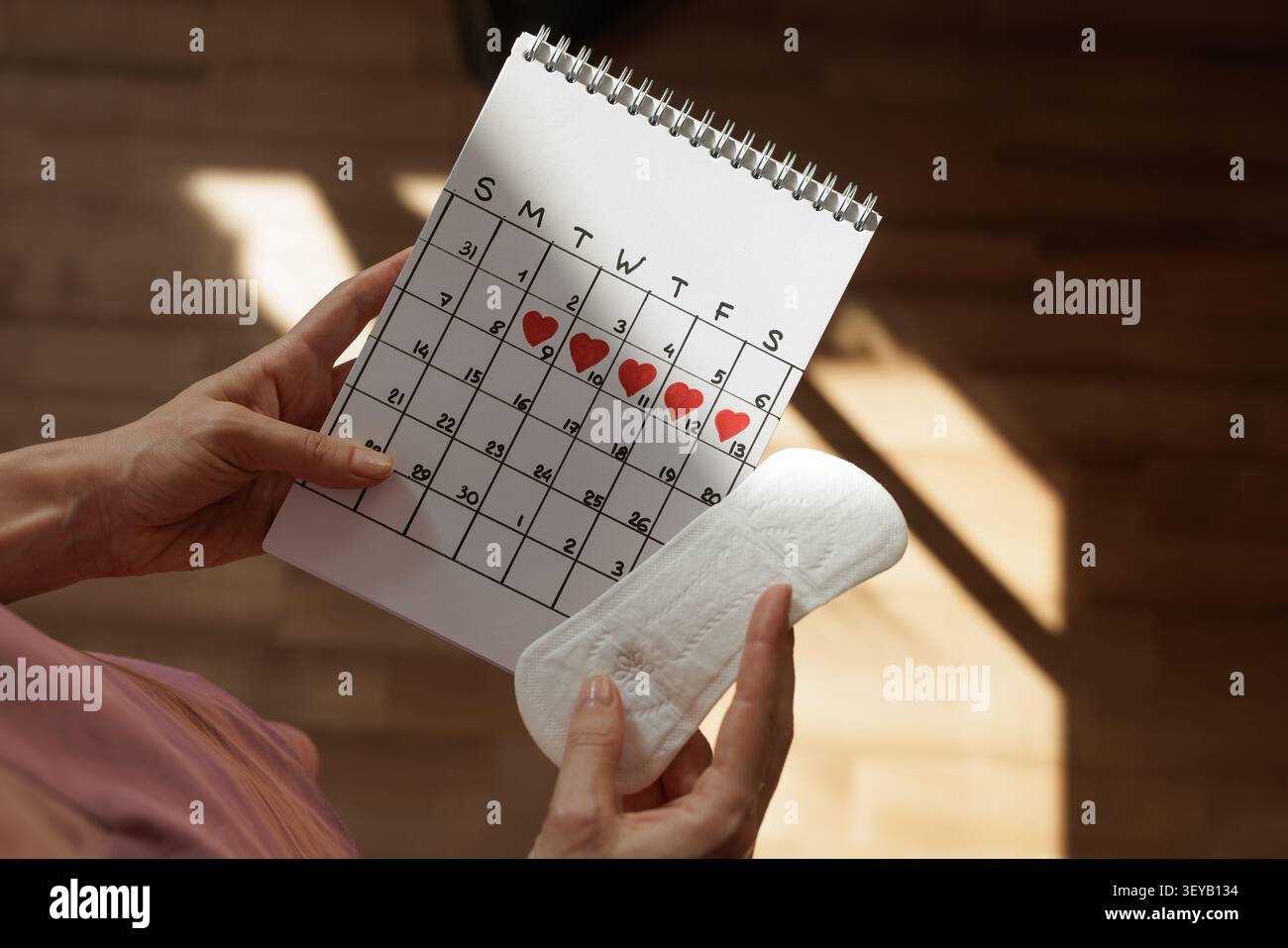 Eine Frau hält einen Kalender mit Herzen, um ihren Menstruationszyklus neben einer Sanitärmatte zu verfolgen, die die Gesundheit der Frauen und die Zyklusüberwachung betont. Stockfoto