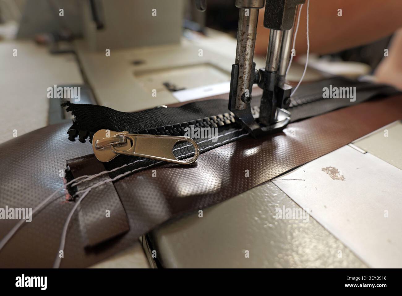 Industrielle Nähmaschine, die einen schwarzen Reißverschluss auf schweres braunes synthetisches Gewebe aufnäht und den Prozess der Textilherstellung und des Handwerkers demonstriert Stockfoto
