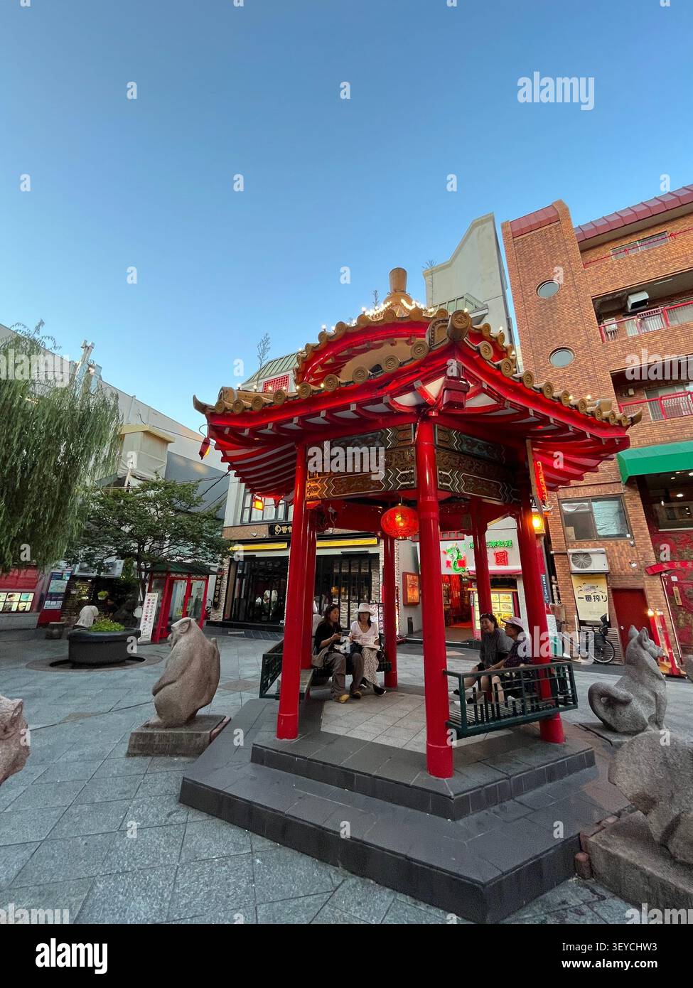 21. Juli 2023, Kobe, Japan - verziertes rotes, chinesisches Pavillon-Tor in Nankinmachi Chinatown mit Löwenstatuen aus Stein und Besuchern in der Abenddämmerung. - Smartphone-aufgenommenes Stockfoto