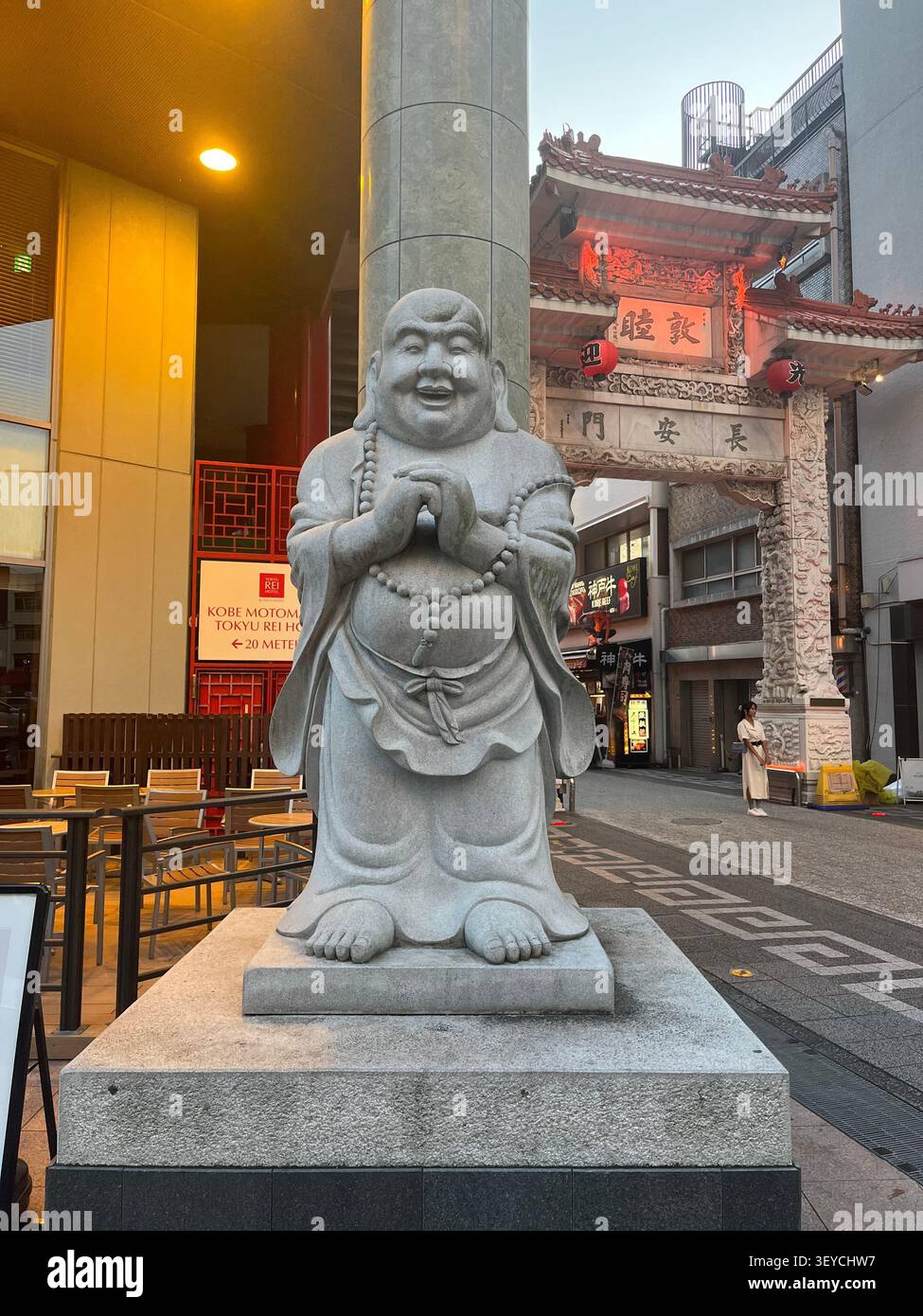 21. Juli 2023, Kobe, Japan - lachender Buddha (Budai/Hotei) Granitstatue am Eingang von Nankinmachi Chinatown, Kobe, mit chinesischem Tempeltor und rotem lan - Smartphone-aufgenommenes Stockfoto