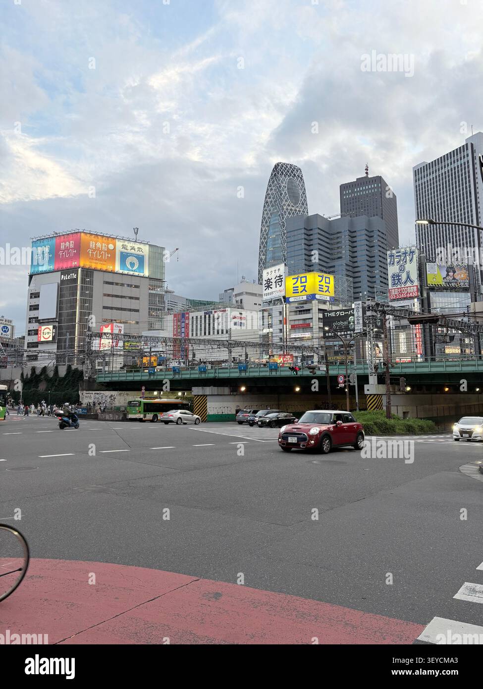 Blick auf die belebte Shinjuku Straße in Tokio, Japan, mit dem ikonischen Mode Gakuen Cocoon Tower, kommerziellen Schildern auf Japanisch und Stadtverkehr. - Smartphone-aufgenommenes Stockfoto