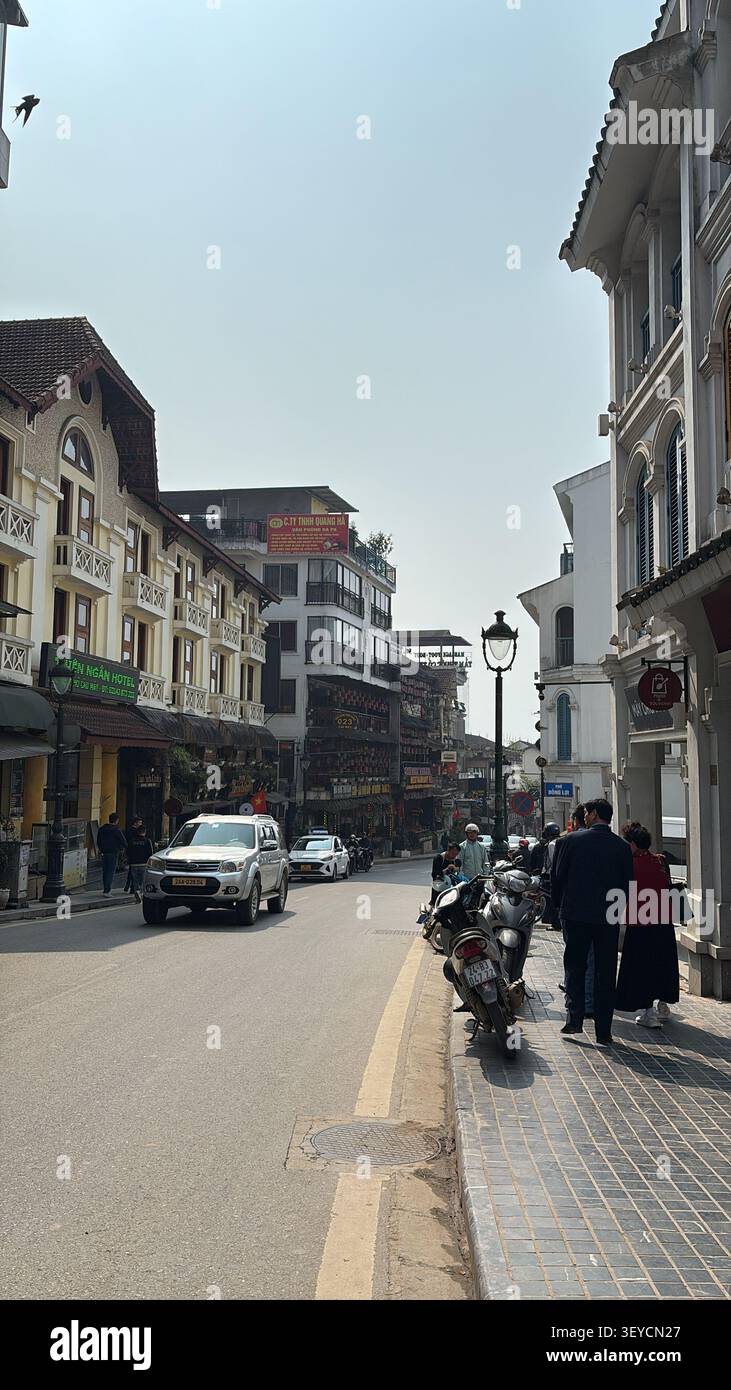 22. März 2025, Sapa, Vietnam - geschäftige Straße in der Altstadt von Sapa mit Gebäuden aus der französischen Kolonialzeit, lokalen Hotels, Motorrädern und Fußgängern an einem trüben Morgen - Smartphone-aufgenommenes Stockfoto