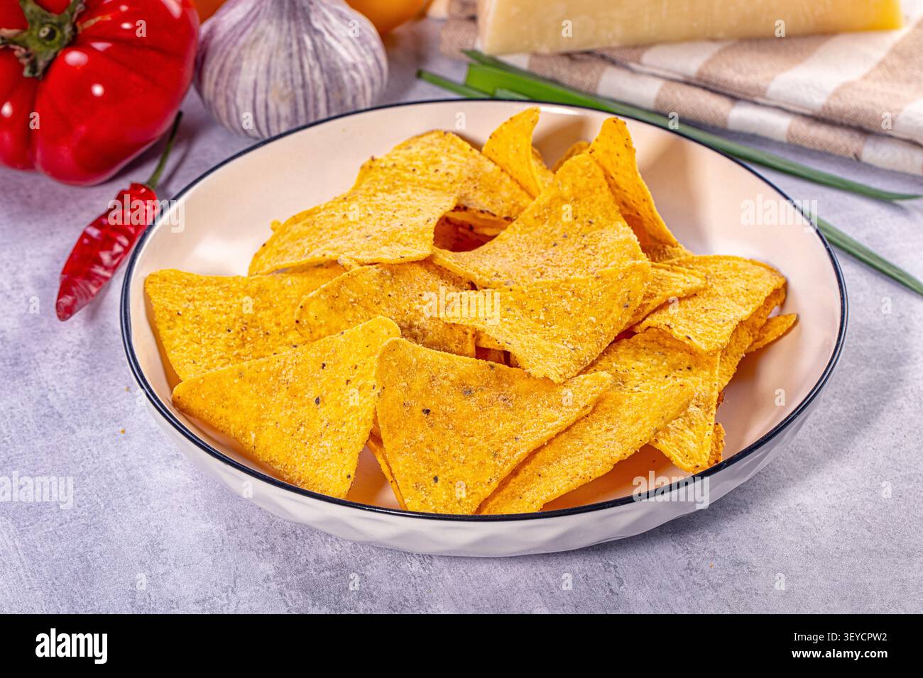Goldene dreieckige Tortillachips in weißer Schüssel mit schwarzem Rand. Hintergrund mit roten Tomaten Knoblauch Chili Pfeffer grüne Zwiebeln Käseblock. Warmes Licht Stockfoto