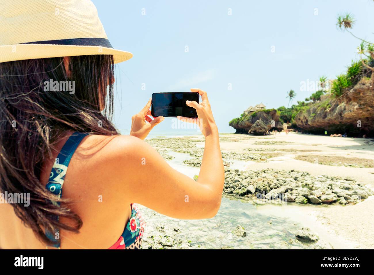 Frau trägt Strohhut verwendet ihr Smartphone, um die dramatischen Korallenklippen und das türkisfarbene Wasser des Mtende Beach in Sansibar zu erfassen. Friedliche Südinsel Stockfoto