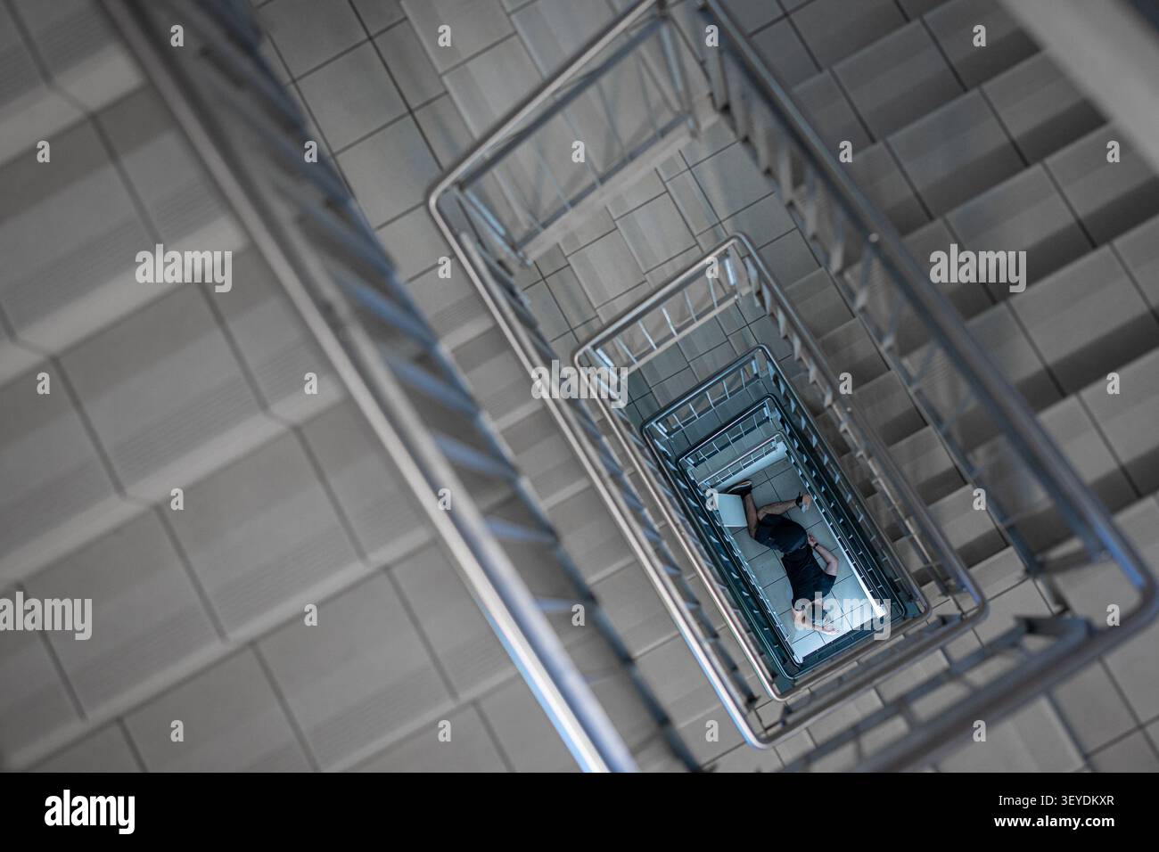 Ein dramatischer Blick von oben auf eine Person, die allein am Fuß einer modernen Treppe liegt. Die geometrische Architektur unterstreicht Themen der Isolation Stockfoto